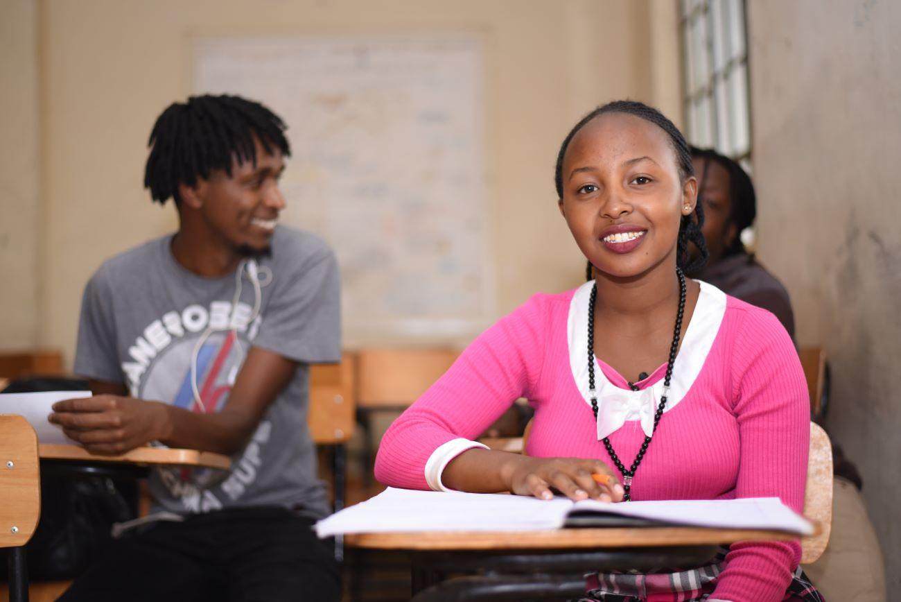 A woman sitting at a desk with a notebook open, ready to study. A man in the background looking at his friend, smiling